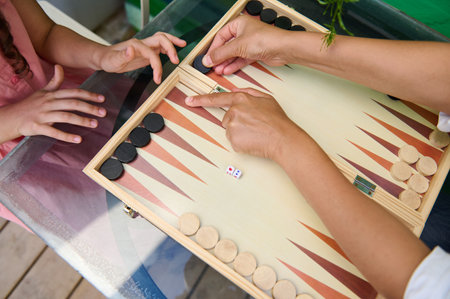 A close-up image of family members enjoying a game of backgammon together, hands moving pieces on...