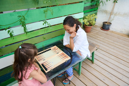 Mother and daughter spending time together playing backgammon, enjoying a sunny day on a patio with...