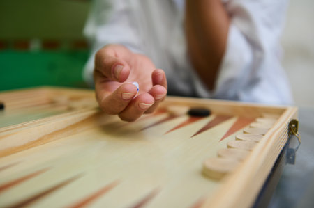 Detailed view of a hand holding a dice while playing backgammon. The setting highlights focus,...