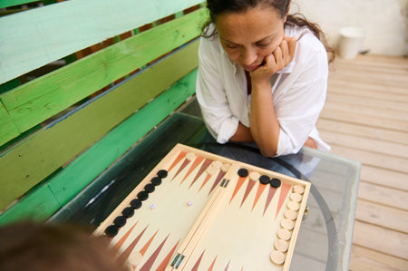 A woman playing backgammon outdoors on a wooden deck, deeply focused on her next strategic move....