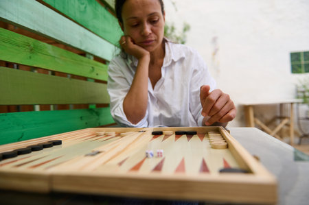 A woman in a white shirt sits outdoors playing backgammon, focused and thoughtful, demonstrating...