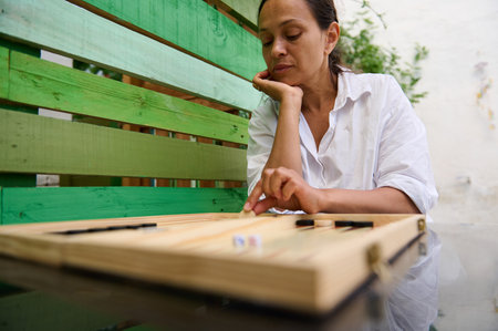A woman sits outside in a garden playing backgammon. She is focused on the board game, enjoying a...