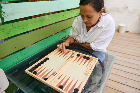 Woman in a white shirt playing backgammon on a glass table in an outdoor setting. She is focused and...
