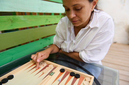 A woman in a white shirt is contemplating her next move while playing a game of backgammon outdoors....