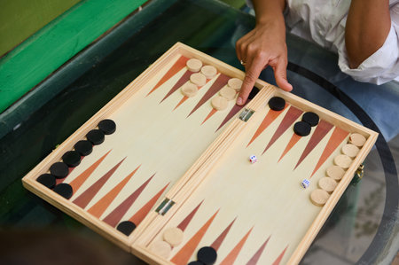 A close-up image showing people playing backgammon on a wooden board, with hands moving pieces and...