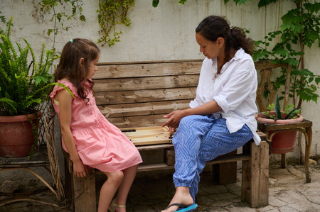 A mother and daughter spending quality time playing a board game together on a wooden bench outside,...