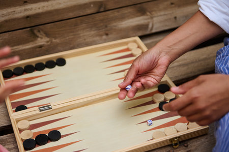 A close-up shot of friends enjoying a game of backgammon on a wooden table outdoors. The image captures the hands of individuals rolling dice and moving pieces.の写真素材