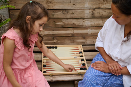 Mother and daughter enjoy a quality time playing a board game outdoors on a wooden bench. Capturing...