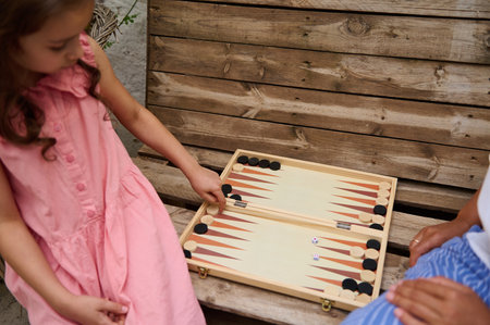 A young girl in a pink dress and an adult play backgammon outdoors on a wooden bench, enjoying...
