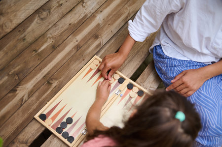 Top view of a parent and child playing a game of backgammon together outdoors. They are seated on a...