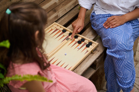 Two people having fun playing backgammon outside on a rustic wooden bench, highlighting family time,...