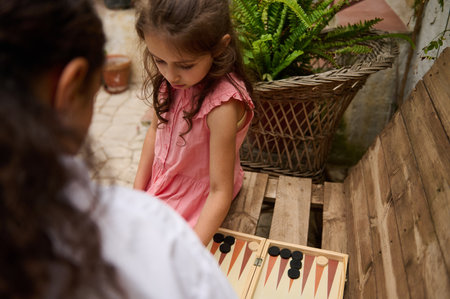 Young girl in a pink dress concentrating on a board game with a friend in an outdoor setting....