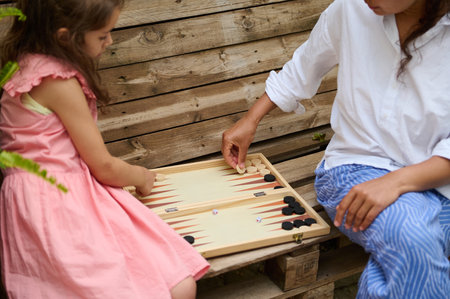A mother and daughter enjoy playing backgammon together outdoors. The young girl in a pink dress and...