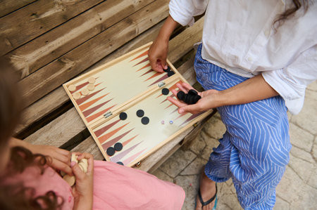 A mother and daughter enjoy a friendly game of backgammon on a wooden bench outside, creating...