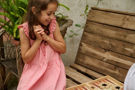 A young girl in a pink dress enjoying a board game while sitting outdoors on a wooden bench. She...