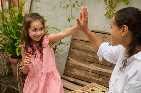 Joyful moment between a young girl and her mother as they high-five during a board game in an...