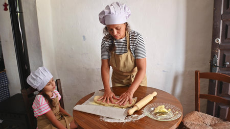 A mother and daughter baking together in the kitchen, kneading dough while wearing chef hats and...