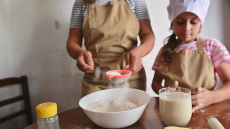 Mother and daughter baking at home, bonding in the kitchen while sifting flour and preparing...