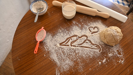 Flour scattered with heart and mountain drawings, surrounded by baking tools and dough on a wooden table, creating a cozy and creative kitchen atmosphere.の写真素材