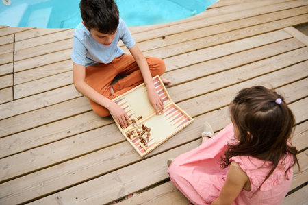 Two kids enjoying a game of backgammon outside next to a pool, sitting on a wooden deck. Relaxed and fun summer activity.の写真素材