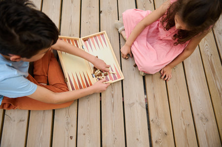 Two children playing a board game on a wooden deck. The image captures a top view of the game in...