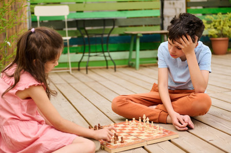 Young boy and girl engaged in a chess game while sitting on a wooden deck in a backyard, showing concentration and strategy.の写真素材