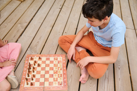 Two children engaged in a chess game on a wooden deck. The image highlights strategy, concentration,...