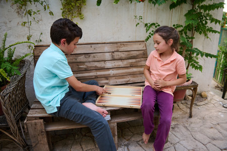 Kids enjoy a game of backgammon outdoors on a wooden bench, surrounded by greenery. Perfect capture...