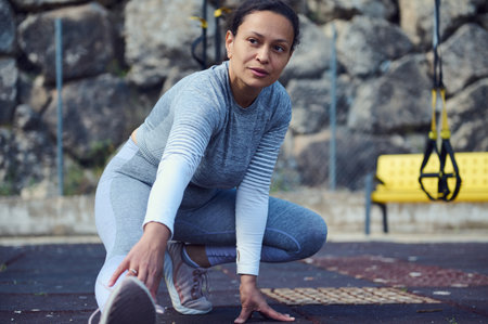 A young woman in sporty attire performs a stretching exercise outdoors. This image captures the...