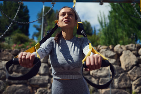 A determined woman exercises with fitness straps outdoors in a sunny park, focusing on building...