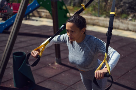 Woman engaging in outdoor fitness using suspension straps in a park, focusing on strength training...