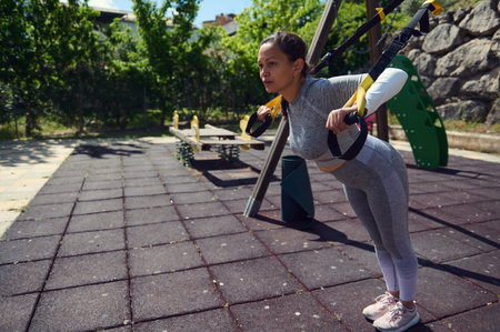 Focused young woman using suspension trainer for a full-body workout in an outdoor park. Emphasizing...