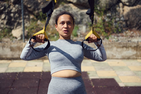 Fit woman using TRX suspension ropes in outdoor workout. Focused on strength and fitness. A sunny...