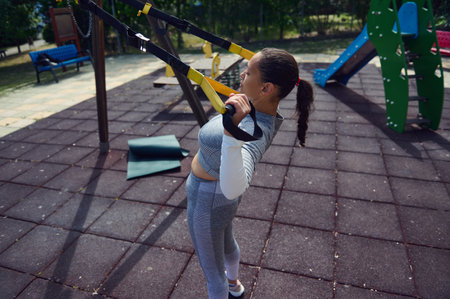 A woman works out with a TRX suspension trainer in an outdoor gym. She demonstrates strength and...