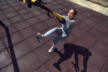 Focused woman using suspension straps for an outdoor workout. She exercises in a park on a sunny...