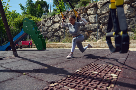A young woman exercises with TRX suspension straps in a park, focusing on strength and fitness in...