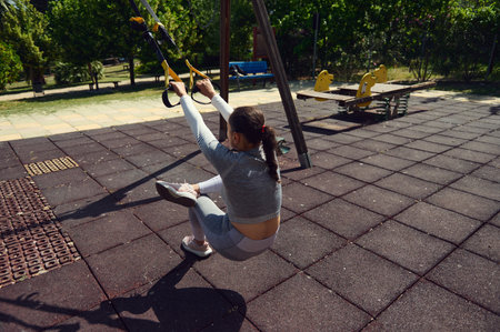 A woman engages in outdoor suspension training in a sunlit park, demonstrating balance and strength....