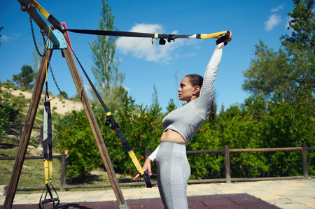 Woman doing outdoor training with a suspension trainer in a park. Focusing on strength and...