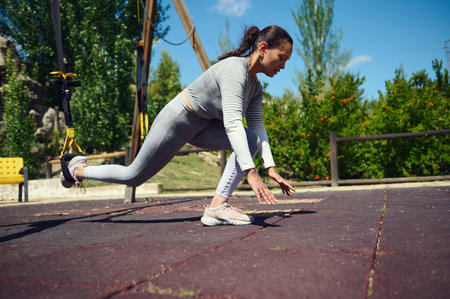 Focused woman performing TRX exercise in the park, enhancing strength and balance. Outdoor workout...