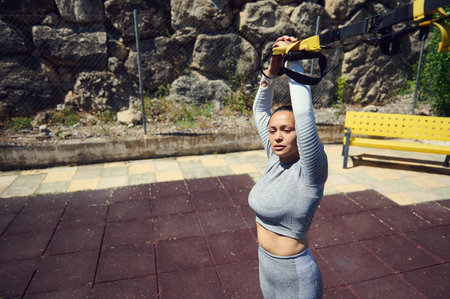 A confident young woman working out with a suspension trainer in an urban park. Fit and focused, she...