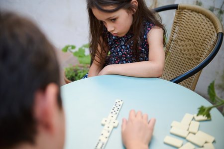 A young girl deep in thought as she plays a game of dominoes, sitting at a table with a friend,...