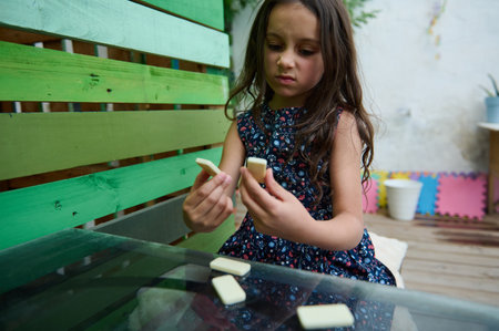 A thoughtful young girl in a floral dress examines domino pieces at a lively, outdoor play area. The...
