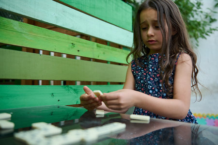 A young girl in a floral dress plays dominoes outdoors, deeply absorbed in thought as she considers her next strategic move.の写真素材
