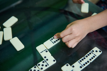 A child's hand strategically places a domino tile on a glass table during a game. The image captures...
