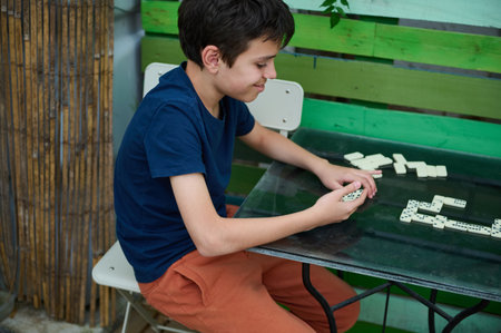 A young boy sits at a colorful table outdoors, engaged in a game of dominoes, concentrated on the...