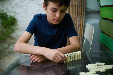 A young boy in a blue shirt concentrates on arranging dominoes on a glass table, surrounded by...