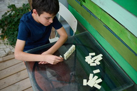A boy intently playing with dominoes on a table outside, emphasizing focus and strategic thinking in...