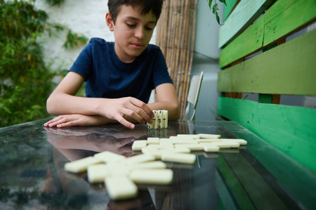 A young boy is focused on playing dominoes in an outdoor setting, emphasizing concentration and...