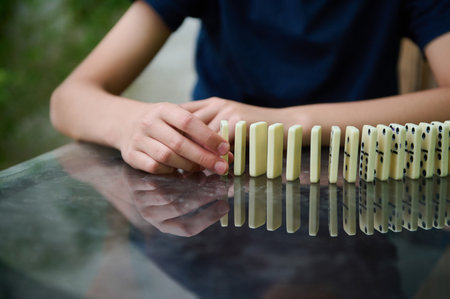 A hand carefully positioned to start a domino chain reaction on a reflective glass table,...