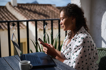 A woman sitting outside, using a smartphone and laptop, enjoying a sunny day with a cup of coffee. Ideal for remote work and lifestyle themes.の写真素材